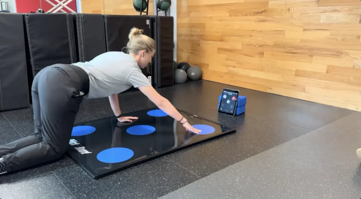 A patient performing the Knee On Ground Plank Iso exercise on a QuickBoard sensor board, with the iPad app on a stand beside it